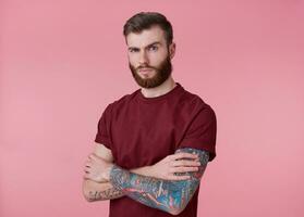 retrato de joven hermoso ceñudo rojo barbado hombre en blanco camiseta, soportes terminado rosado antecedentes con cruzado brazos, mira a el cámara con malentendido. foto