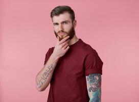 Portrait of young attractive thinking tattooed red bearded man in red t-shirt, looks at the camera and touches the chin, stands over pink background. photo