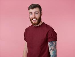 Young handsome happy amazed red bearded man in blank t-shirt, stands over pink background, looks surprised at the camera with wide open mouth and eyes. photo