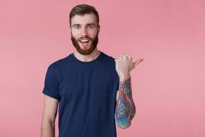 Prttrait of young happy amazed red-bearded young guy ,with wide open mouth in surprise, wearing a blue t-shirt, pointing finger to copy space at the right side isolated over pink background. photo