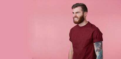 Portrait of young handsome evil red bearded man in blank t-shirt, looks aggressive and shocked, stands over pink background, ooks to copy space on the left side. photo