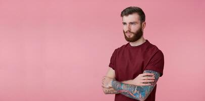 retrato de joven hermoso alegre rojo barbado tatuado hombre en rojo camiseta, mira contento y sonrisas, mira lejos, soportes terminado rosado antecedentes con copyspace a el izquierda lado. foto