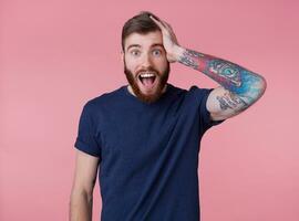 Portrait of young happy amazed attractive red-bearded young guy , wearing a blue t-shirt, with wide open mouth in surprise, hold on to head, heard very good news, isolated over pink background. photo