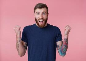 Young happy red-bearded guy with blue eyes, wide open mouth with delight, celebrates victory of his favorite football team's , with arms raised up and clenched fists, isolated over pink background. photo