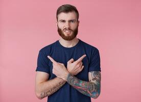 Young red-bearded frowning guy , deceived, fingers crossed, fingers pointing in different directions, isolated over pink background. photo