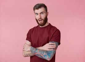 retrato de joven tatuado preguntado rojo barbado hombre en blanco camiseta, soportes con cruzado brazos terminado rosado fondo, mira a el cámara con abierto boca. foto