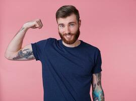 Portrait of young red-bearded handsome manful guy demonstrates biceps and strength, pleased with himself, broadly smiling and looking at the camera isolated over pink background. photo