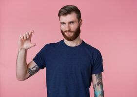 Portrait of young attractive red-bearded guy, frowning and looking at the camera, shows fingers something small with an insidious grin slyly looking at the camera isolated over pink background. photo