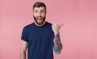 Young happy red-bearded young guy , with wide open mouth in surprise, saw something wrong, wants to draw your attention pointing finger to copy space at the right side isolated over pink background. photo