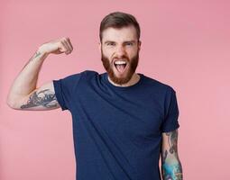 Young screaming red-bearded handsome manful guy demonstrates biceps and strength, looking at the camera isolated over pink background. photo