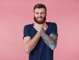 Portrait of young red-bearded unhappy man, can't find a way out, displeased, trying to strangle himself, scowl and grin isolated over pink background. photo