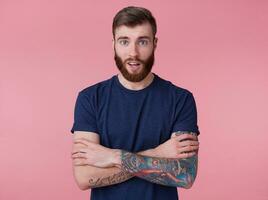 Portrait of young happy amazed attractive red-bearded young guy with crossed arms, wearing a blue t-shirt, looking at the camera with wide open mouth in surprise isolated over pink background. photo