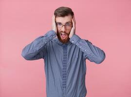 Shocked young handsome red bearded man with glasses and a striped shirt, put his head in his hands, stands with wide open mouth over pink background. photo