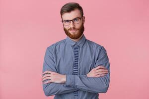 retrato de un grave estricto barbado joven hombre usa un a rayas camisa, siente enojado y mantiene su brazos cruzado aislado en un rosado antecedentes. foto