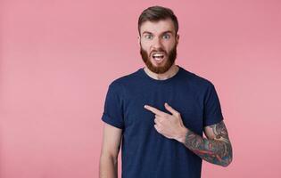 joven atractivo de barba roja chico con azul ojos mira indignado, vistiendo un azul camiseta, ceñudo con amplio abierto boca, señalando dedo a copias paso a el izquierda lado aislado terminado rosado antecedentes. foto