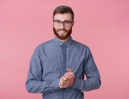 Young attractive red-bearded young guy with glasses and a striped shirt smiles broadly, folded his arms and looking affably at the camera, stands over pink background. photo