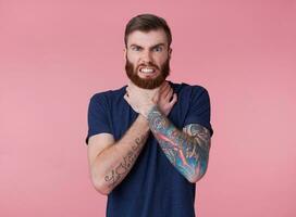 Young red-bearded frowning guy, can't find a way out, displeased and angry, trying to strangle himself, scowl and grin isolated over pink background. photo