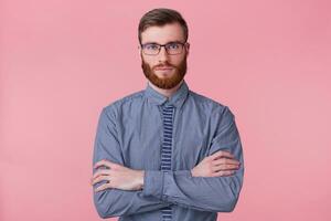 Portrait of a calm bearded young man wears a striped shirt, keeps his arms crossed and looking at the camera isolated on a pink background. photo