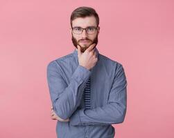 Portrait of young attractive red-bearded young guy with glasses and a striped shirt, touches his chin, looks thoughtfully at the camera with calm expression on his face stands over pink background. photo