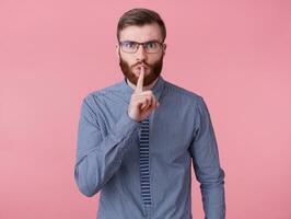 Young handsome red bearded man with glasses and a striped shirt, keeps finger on lips, tells secret information, demonstrates hush gesture isolated over pink background. photo