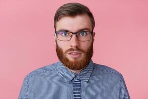 Close up of young attractive bearded man in a striped shirt with glasses, looks at the camera in surprise, does not believe his eyes, isolated over pink background. photo