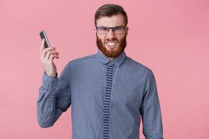 Photo of a young handsome bearded man with glasses and a striped shirt, holding the phone away from his ear, because he is called by a furious boss who kits a phone. Isolated over pink background.