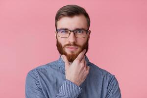 Photo of serious young attractive bearded man in a striped shirt with glasses, holds hand near the chin, looking camera ponder about something, isolated over pink background.