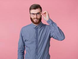 Displeased handsome young handsome red bearded man with glasses and a striped shirt, stands over pink background, raising his eyebrow questioningly, doubts and looks at the camera. photo
