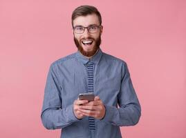 Young attractive red-bearded man with glasses and a striped shirt smiles broadly, holding a phone and enjoys great wi fay, stands over pink background. photo