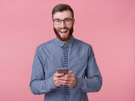 joven atractivo de barba roja hombre con lentes y un a rayas camisa sonrisas en general, participación un teléfono y disfruta genial Wisconsin meter, soportes terminado rosado antecedentes. foto