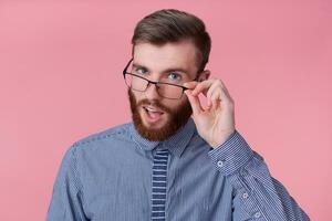 joven alegre atractivo de barba roja joven chico con lentes y un a rayas camisa, mira a el cámara mediante lentes y sonrisas en general, soportes terminado rosado antecedentes. foto