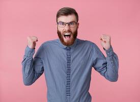 Portrait of displeased angry young handsome red bearded man with glasses and a striped shirt, stands over pink background, screaming with fists raised. photo