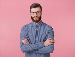 Displeased young handsome red bearded man frowns face, looks at the camera with disgust, with crossed arms stands over pink background. photo