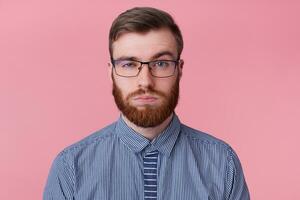 Close up of serious young attractive bearded man in a striped shirt with glasses, one eyebrow raised, looking at the camera isolated over pink background. photo