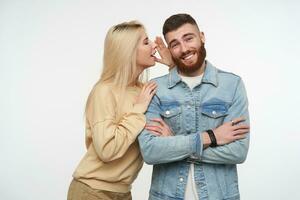 Cheerful young bearded brunette lovely guy crossing hands on his chest while listening to his pretty long haired blonde girlfriend, isolated over white background photo