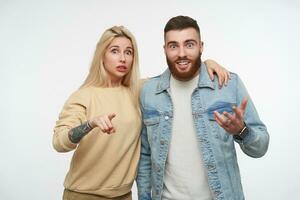 Agitated young pretty couple in casual wear rounding their blue eyes while looking amazedly at camera and raising surprisedly eyebrows, isolated over white background photo