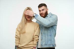 Bewildered young handsome short haired brunette bearded man raising his hand while closing eyes of his blonde surprised girlfriend, isolated over white background photo