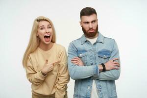 Offended young brunette bearded man folding his hands on chest while pretty long haired blonde female showing excitedly on him with wide mouth opened, isolated over white background photo