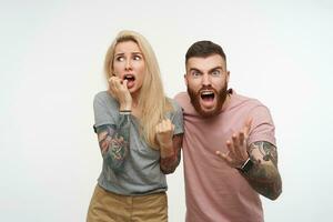Excited young handsome brunette tattooed guy screaming excitedly and raising his hand while posing over white background with scared pretty blonde female in grey t-shirt photo