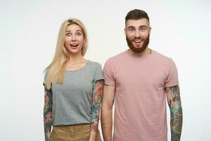 Joyful young pretty couple with tattooes looking surprisedly at camera and smiling broadly, standing over white background and keeping their hands down photo