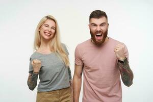 Overjoyed young lovely couple with tattooes rejoicing abouth something and raising emotionally hands, wearing casual t-shirts while standing over white background photo