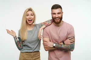 Joyful young lovely couple with tattooes laughing happily while rejoicing about something, wearing casual clothes while being isolated over white background photo