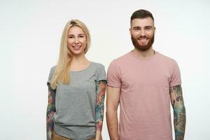 Studio photo of young lovely cheerful tattooed couple looking positively at camera with wide smiles and keeping their hands along bodies, posing over white background