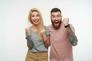 Indoor photo of happy young lovely tattooed couple in casual clothes raising joyfully their fists while rejoicing about something, standing over white background