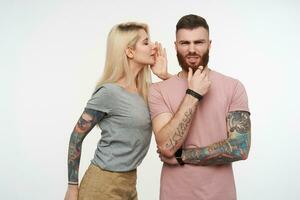Studio photo of young attractive tattooed guy with beard frowning his face while looking pensively at camera and listening to his pretty long haired blonde girlfriend