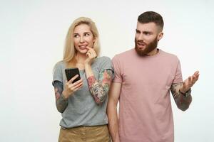 Glad young pretty long haired blonde tattooed woman looking cheerfully aside while holding mobile phone, standing over white background with gloomy bewildered bearded man photo