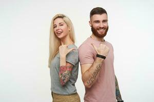 Cheerful young lovely couple with tattooes showing on each other with thumb and looking gladly at camera with wide smiles, standing over white background photo