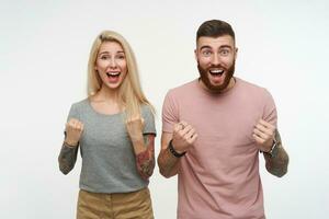 Agitated young tattoed pretty couple in casual clothes raising excitedly hands while looking joyfully at camera with happy faces, standing over white background photo