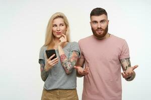 Positive young lovel blonde tattooed woman keeping mobile phone and looking cunningly aside while posing over white background with confused bearded brunette guy photo