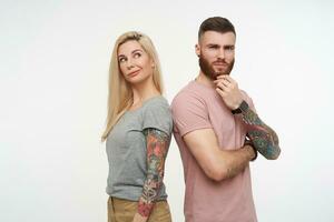 Bewildered pretty pair of young people standing back to back while thinking about something and keeping their lips folded, posing over white background photo